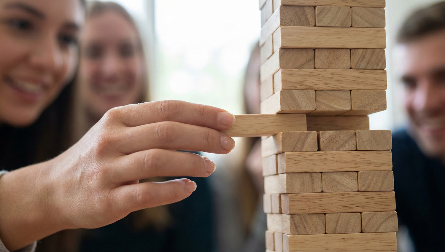 A hand slowly pulls a wooden block from a tower while friends watch and smile in the background.